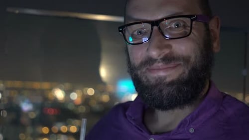 Smiling man enjoying cocktail and smartphone on rooftop terrace at night