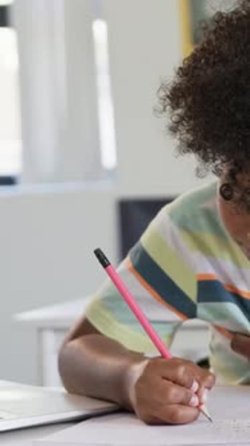 Vertical video: Sitting at desk, child in pastel-striped shirt doing homework with pink pencil
