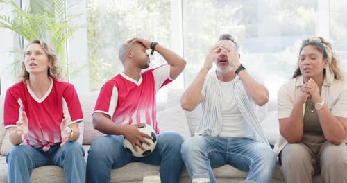 Diverse group on sofa two wearing red, with ball, watching match, reacting to play, reaching snacks