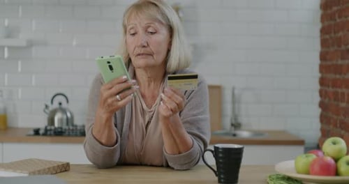 Senior Woman Shopping Online Paying with Credit Card and Smartphone Indoors in Kitchen at Home