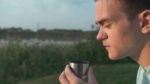A Young Guy Drinks Coffee From A Thermos Behind A Lake