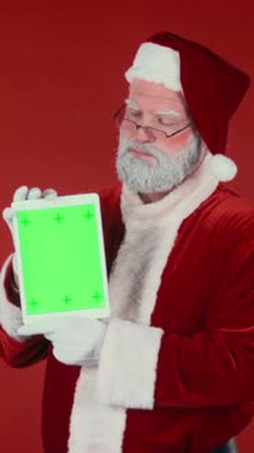 Vertical of Santa Claus Looking at Tablet with Green Screen on Red Background