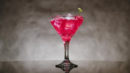 Cocktail in glass with ice decorated with cherries on gray background rotating on table.