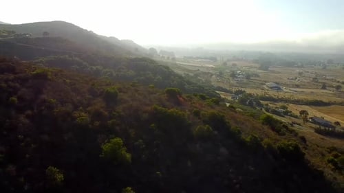 Drohnenschuss über Hügel mit Sonnenschein durch Nebel und Farm im Hintergrund.