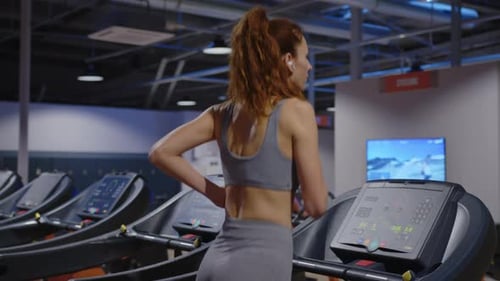 Rear View a Girl with Long Hair in Headphones Runs on a Treadmill in a Modern Gym