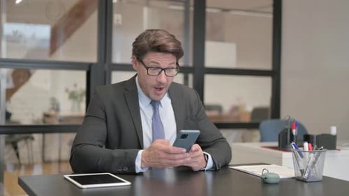 Middle Aged Businessman Celebrating Online Success on Phone while Sitting in Office