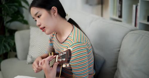 Beautiful young woman plays ukulele and sings in living room at home during holiday.