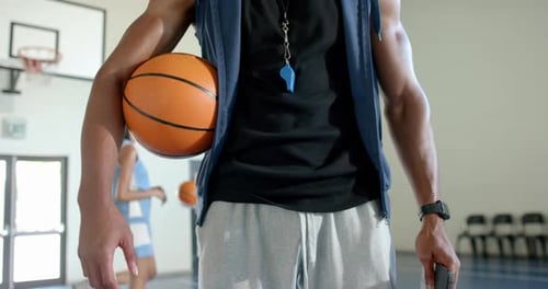 Holding basketball, coach with whistle standing in school gym, players practicing