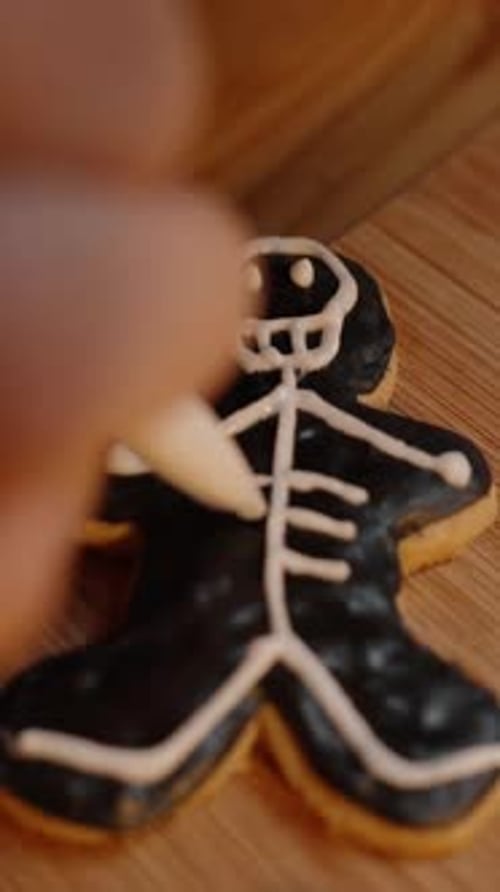 Vertical video. Drawing a Skeleton with White Icing on a Black Gingerbread Man-Shaped Cookie.