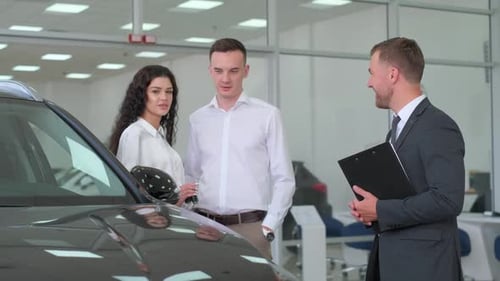 Car Dealership Cheerful Manager Guy Showing Automobile To Customers In Auto Dealership Store