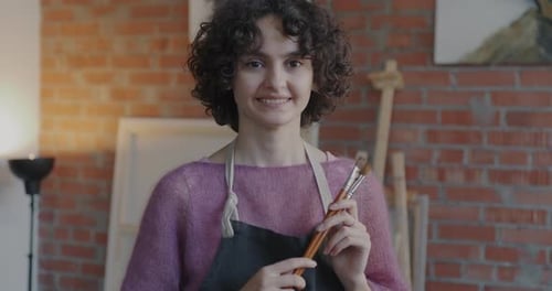 Portrait of Talented Young Woman Artist Standing in Studio Holding Brush Ready for Painting Smiling
