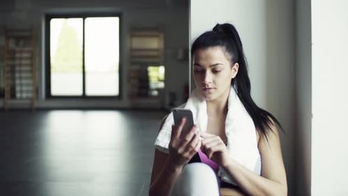 Young woman resting after workout texts on her smartphone in the gym