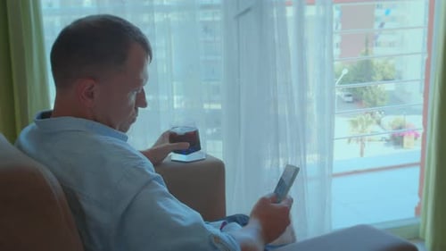 Young Man Pondering By the Window with a Cold Drink and a Phone in Hand Side View Chair