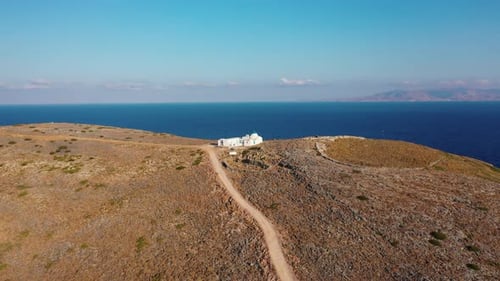 Aerial shot of a dirt road leading to a Greek Monastery on top of a hill by the sea. The cycladic is