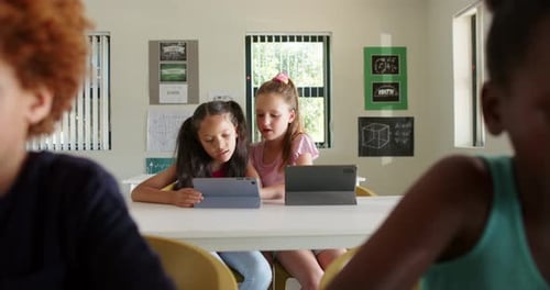 Diverse girls tapping tablet, leaning in and pointing, collaborating in class on math task at table