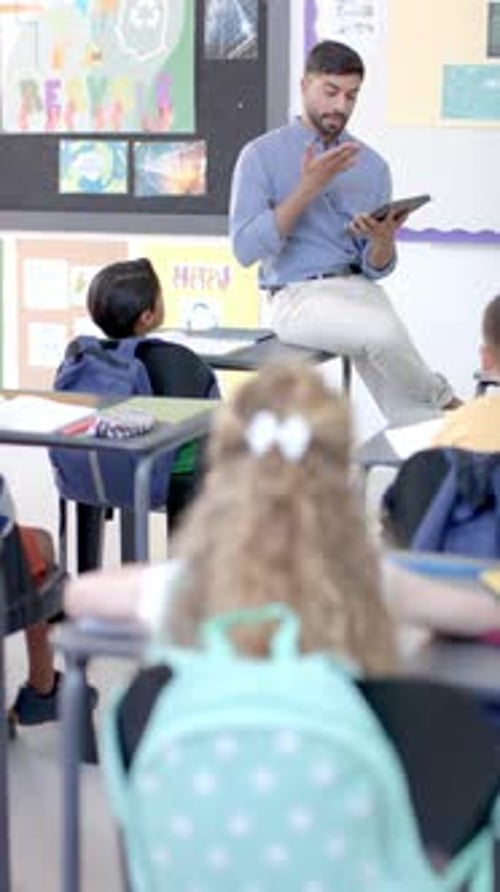 Asian male teacher with short black hair sits on desk, hand raised in vertical video