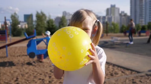 Little Girl Child Inflates a Yellow Balloon in the Summer Park