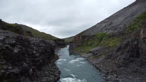 Drone captures river winding through rugged rocky cliffs beneath cloudy sky in Iceland, offering dra