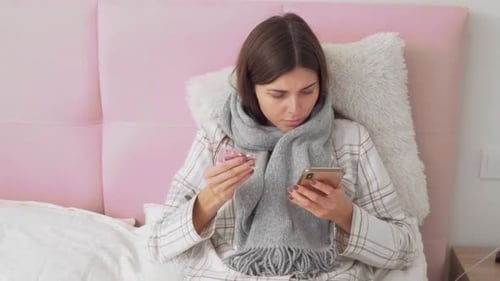 sick girl lying in bed reading on the phone about pills and drinking tea