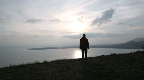 Man Stands on Hilltop Overlooking Lake at Sunset