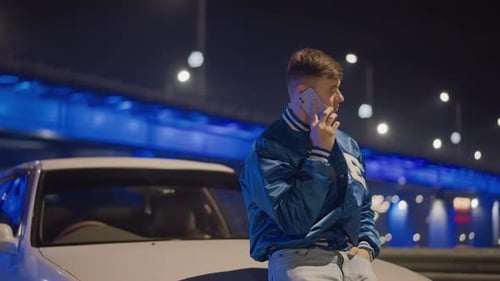 White Man Talking on Phone Under Bridge Leaning on Car Hood Neon Lights and Traffic Bokeh Anxious