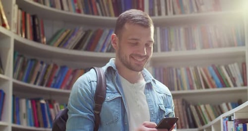 Young Caucasian Male Student with Backpack Chatting on Smartphone and Sending Messages in Library