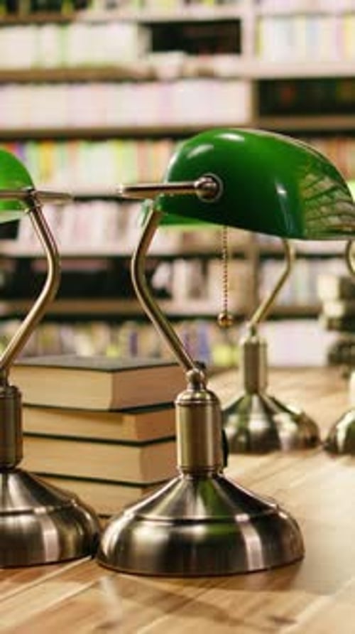 Vertical Video Stack of Books Placed on Wooden Library Desk Beneath Green Lamps