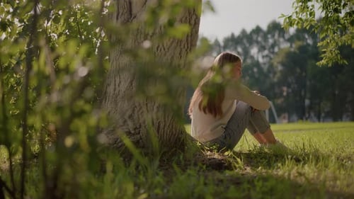 Thoughtful Woman Sitting on Grass in Sunlit Park Surrounded By Greenery