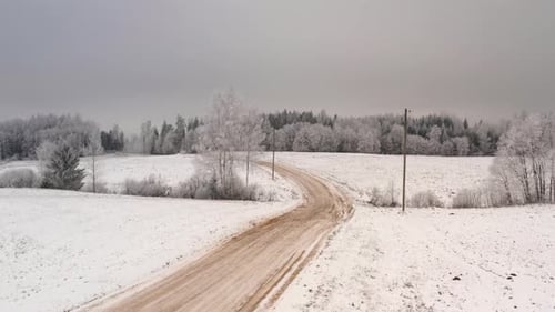 Flying over gravel road in countryside. Aerial winter day scenery with forest covered in snow and fr