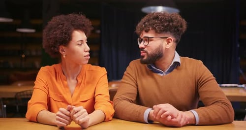 Homme et femme confiants assis à table parlant et faisant des gestes tout en regardant directement la caméra