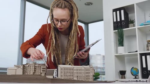 Woman with dreadlocks in glasses working with model of future buildings in beautifully designated de