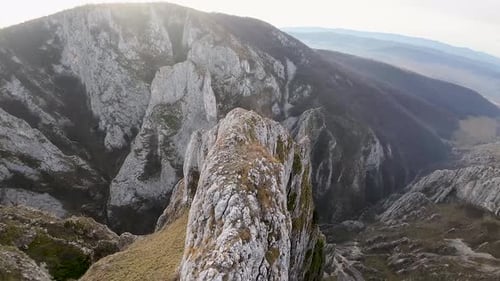 Aerial zoom-out shot showcasing a rugged mountain cliff with a deep valley below, emphasizing the dr