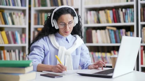 Female student in headphones listens to online e-learning, video course in campus library space