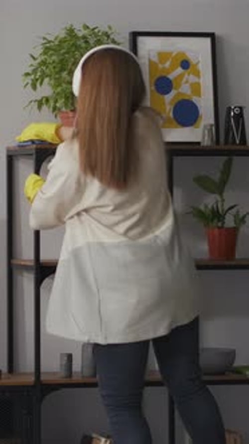 Woman Wearing Headphones and Rubber Gloves Dusting Shelves and Arranging Potted Plants in a Modern