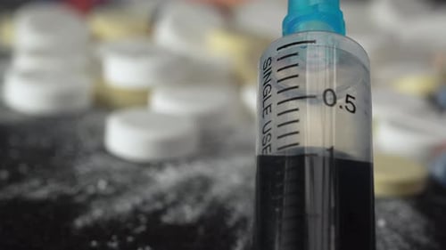 Close-up syringe plunger with blue dangerous liquid with blurred background of pills and medicines