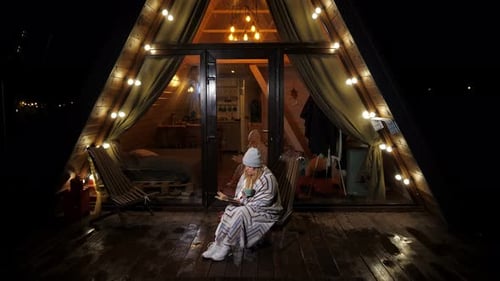 A Woman Wrapped in a Blanket is Sitting Reading a Book Near a Triangular House