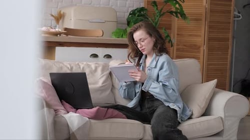 Woman on Couch Writing in Notebook with Laptop