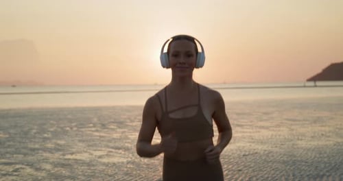 Portrait of Pretty Woman with Earphones Runs and Smiles on Sea Beach at Sunset