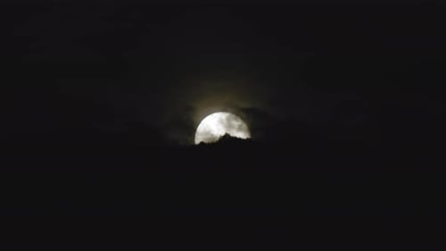 The Full Moon Rises in the Night Sky Behind Trees on a Hill as Dark Clouds Pass by
