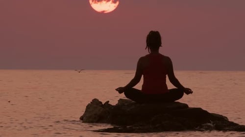 Woman meditates while sitting on stone in water of sea or ocean on background of amazing sunset sky