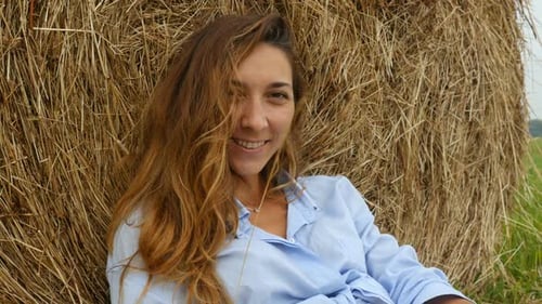 Pretty Young Woman Stands Near Haystack And Plays With Straw. Farmer's Wife Walks On The Field