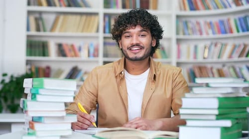 Portrait of smiling college student studying sitting at desk taking notes in campus library space