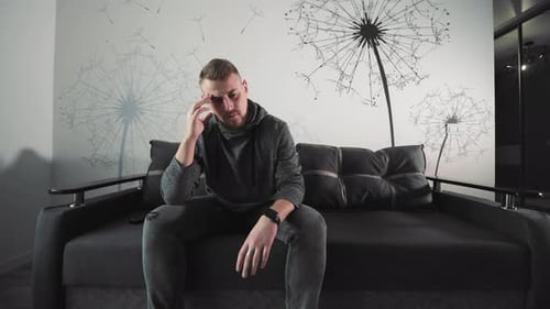 A Depressed Man is Sitting on a Sofa A Pensive Man Sits on a Grey Sofa in a Stylish Room Lost in