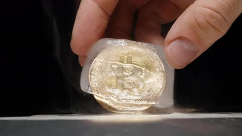 Hand Holding a Bitcoin Coin Encased in a Partially Melted Ice Cube on a Heated Metal Surface with