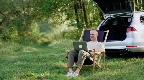 Smiling male freelancer work remote on laptop. Man in glasses is happy to work in nature.
