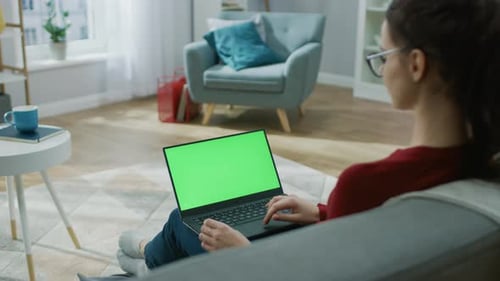 Young Woman at Home Works on a Laptop Computer with Green Mock-up Screen. She's Sitting On a Couch