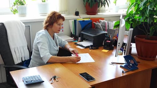 Woman Working at a Cluttered Office Desk with Folders Documents and Plants in the Background Concept