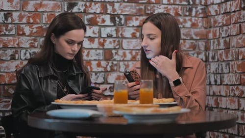 Two Beautiful Girls are Sitting in a Cafe and Talking with Phones