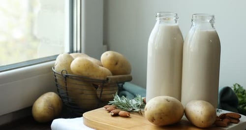 Leche vegana en botellas de vidrio, patatas, romero y almendras en una mesa de madera cerca de la ventana, primer plano