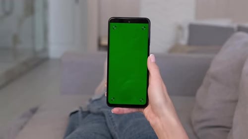 A Woman Makes a Swipe Down Gesture on the Green Screen of a Smartphone While Lying on the Sofa at